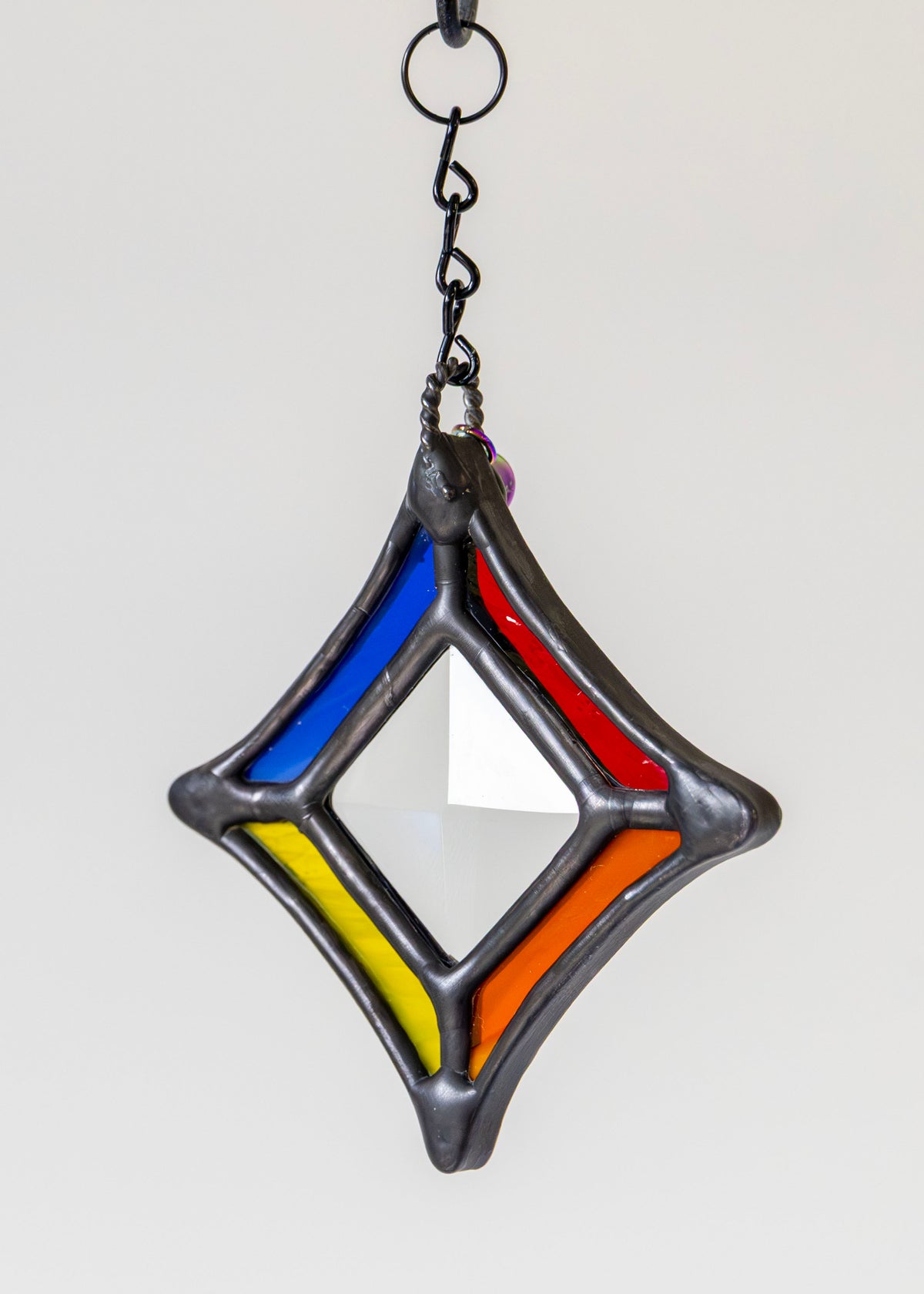 A hanging stained-glass style ornament or suncatcher with a diamond-shaped clear glass center surrounded by colored panels in blue, red, yellow, and orange, outlined in dark gray metal.