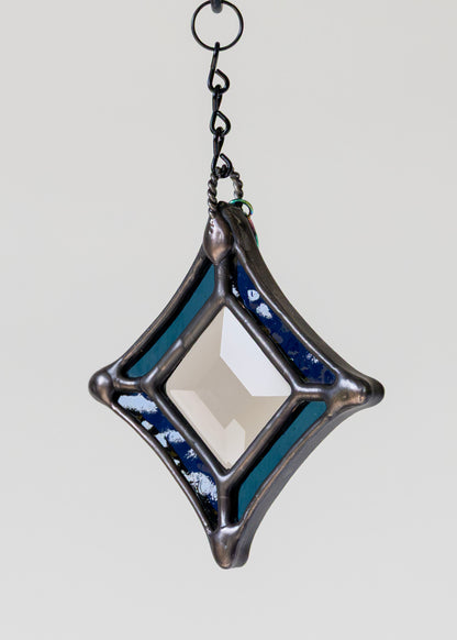 A diamond-shaped stained glass window panel with a central clear glass section, surrounded by textured blue and teal glass sections and a dark metal frame, hanging from a black chain against a light gray background.